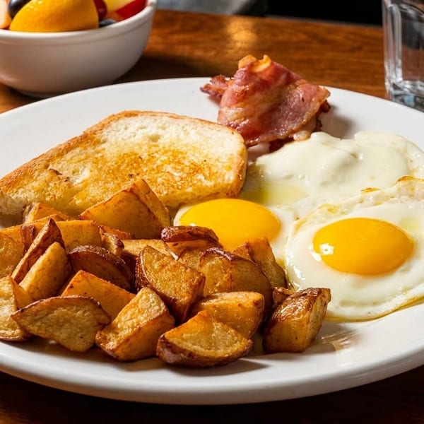 A full breakfast plate with fried eggs, crispy home fries, bacon, and toast, with a fruit cup.