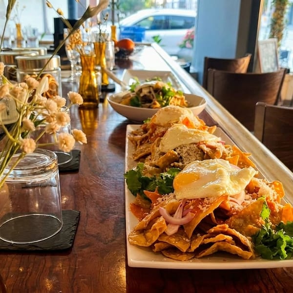A long plate of chilaquiles with fried eggs, a cocktail, and a salad on a wooden bar.