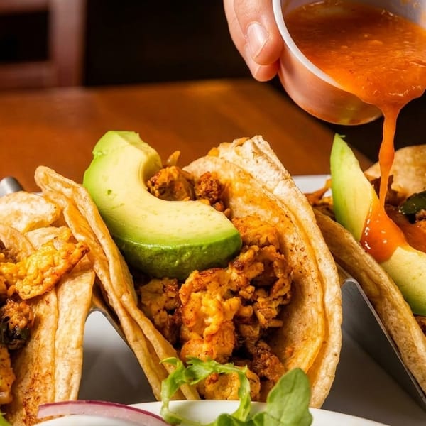 Three tacos with meat and avocado slices, with salsa being poured onto one.