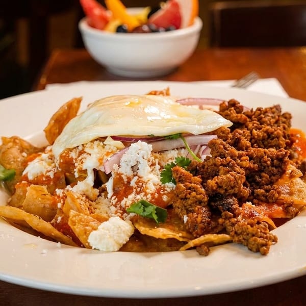 Chilaquiles with a fried egg, chorizo, cheese, and red onions, with a fruit bowl and coffee.