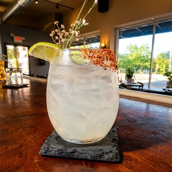 A refreshing cocktail with a chili-lime rim and lime wedge sits on a dark wooden bar.